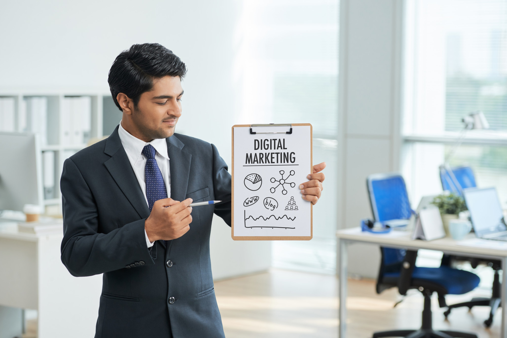 "Business professional in a suit holding a clipboard labeled 'Digital Marketing' with hand-drawn icons, representing strategies in a modern office setting."